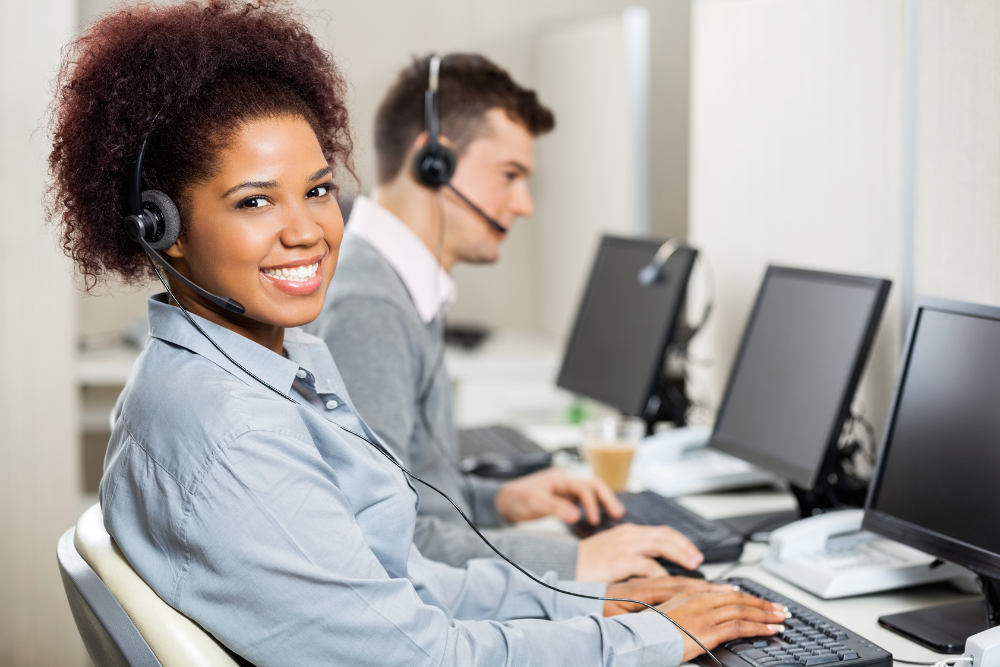 Customer service staff wear headsets and work at computers, one smiling toward the camera.