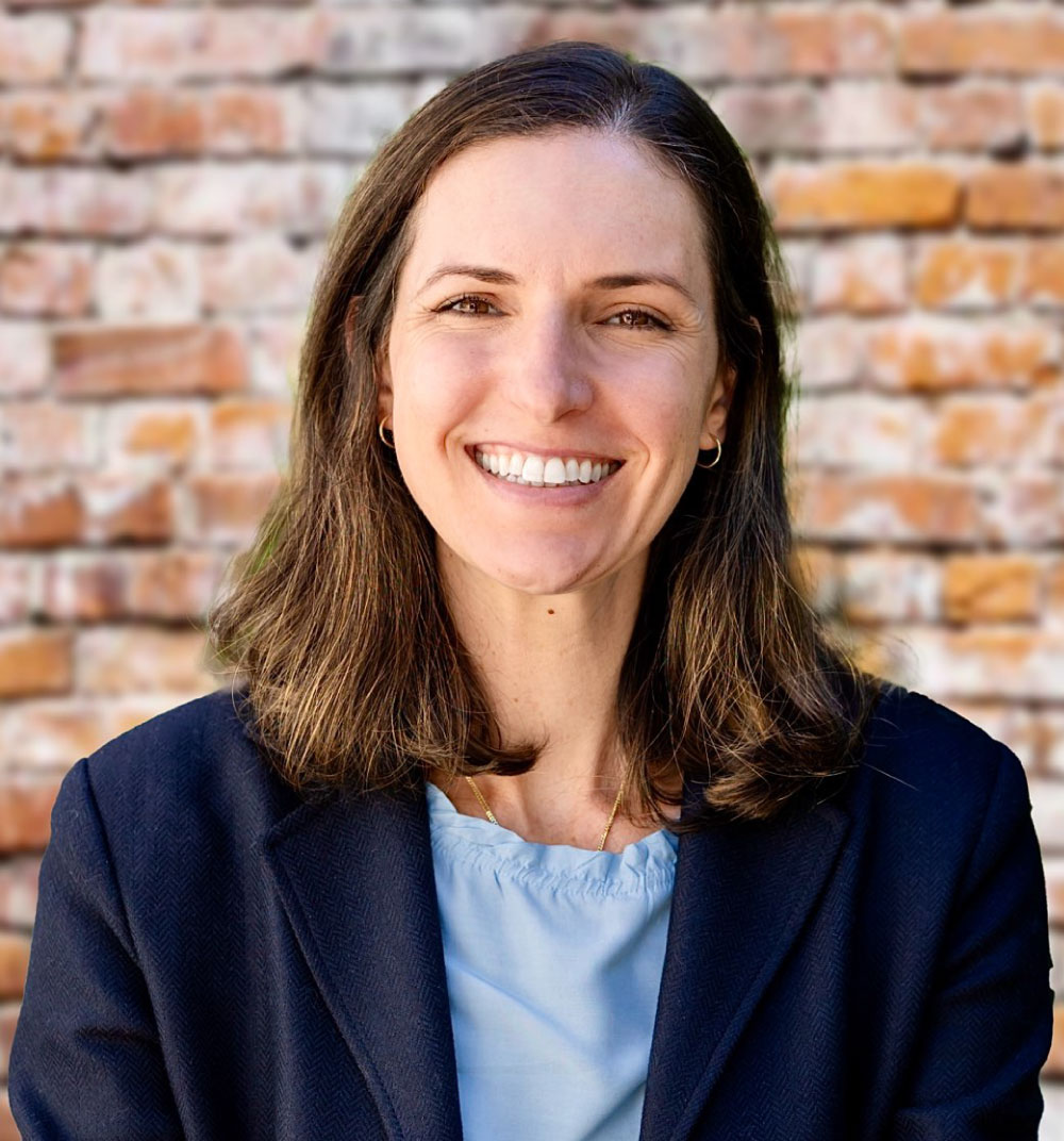 A woman wearing a blue shirt and black blazer smiles warmly at the camera.