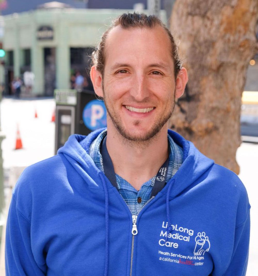 A man in a blue sweatshirt smiles warmly at the camera, exuding a friendly and approachable demeanor.