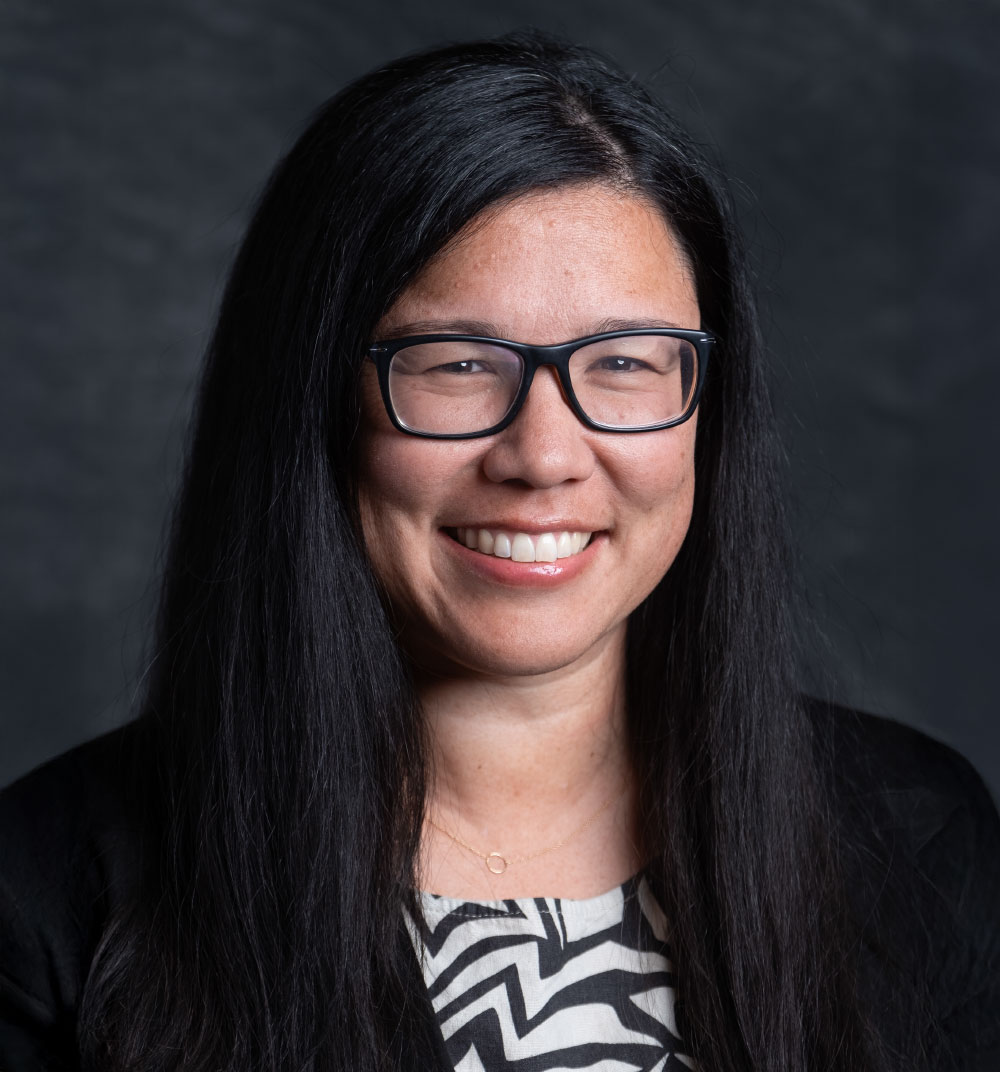 A woman with long black hair and glasses smiles warmly at the camera.