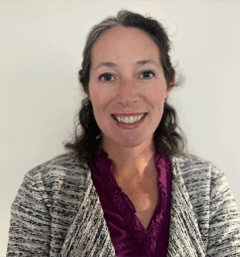 A woman wearing a purple shirt and cardigan jacket stands confidently, smiling at the camera.