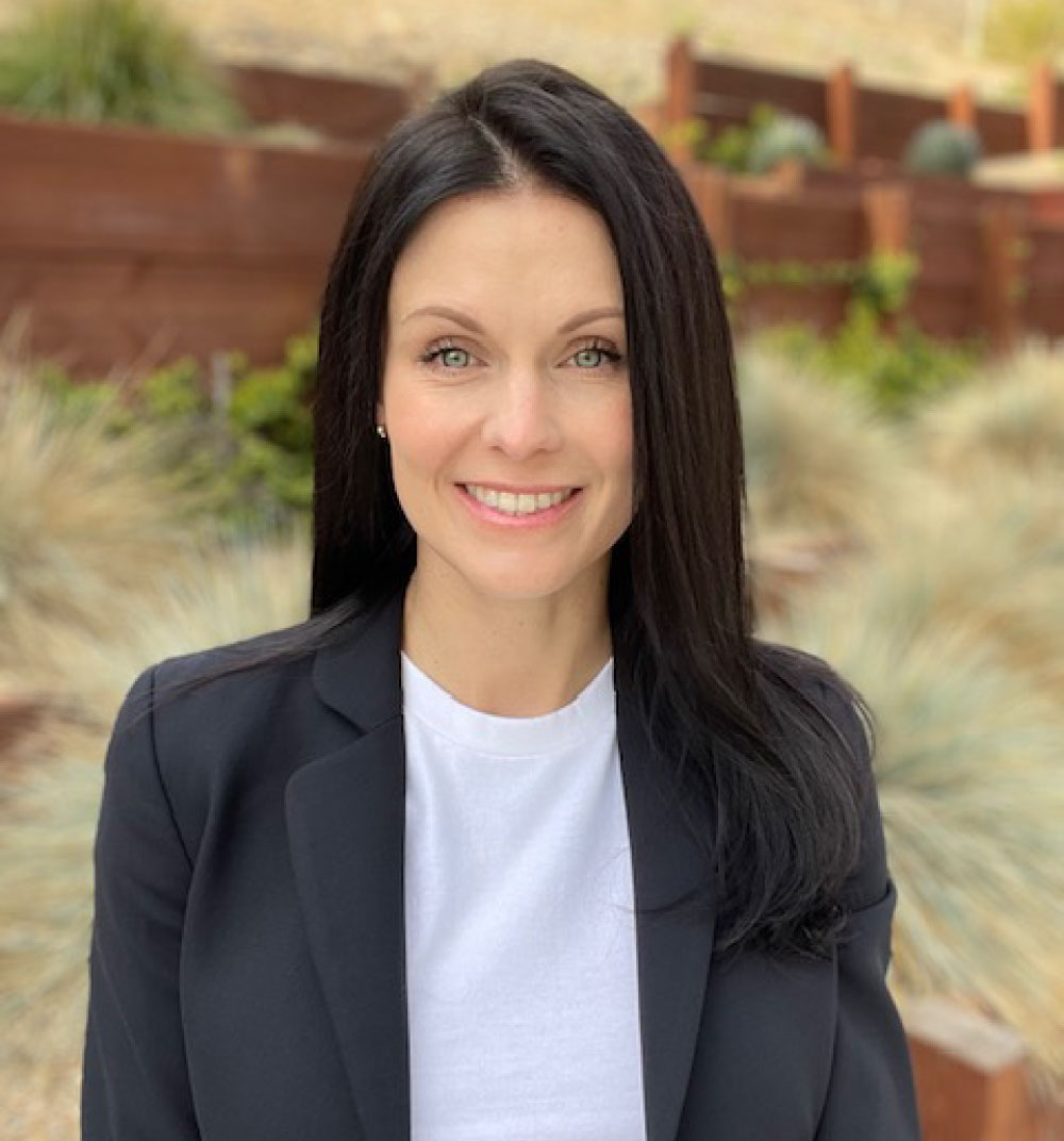 A woman with long black hair wearing a white shirt stands confidently, smiling at the camera.
