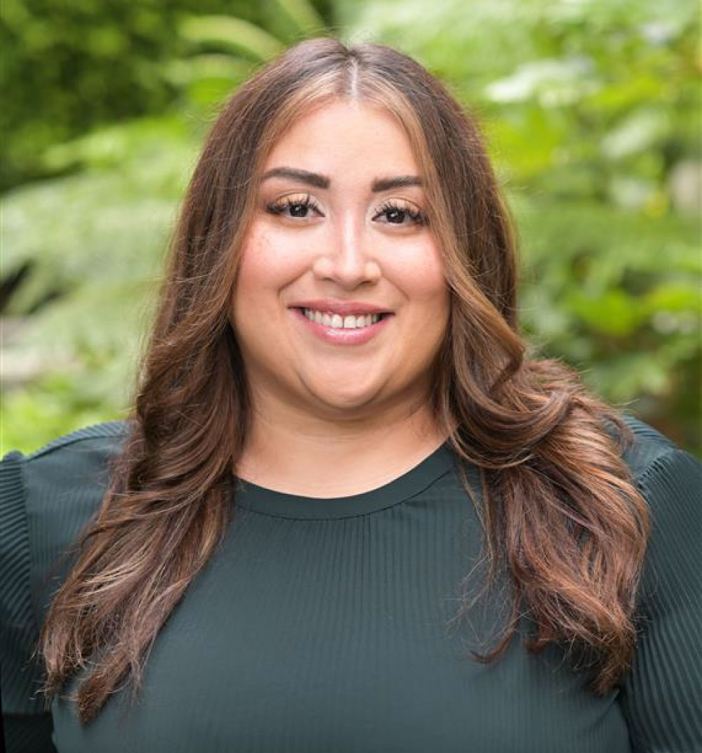 A woman with long brown hair wearing a green shirt stands, smiling at the camera.