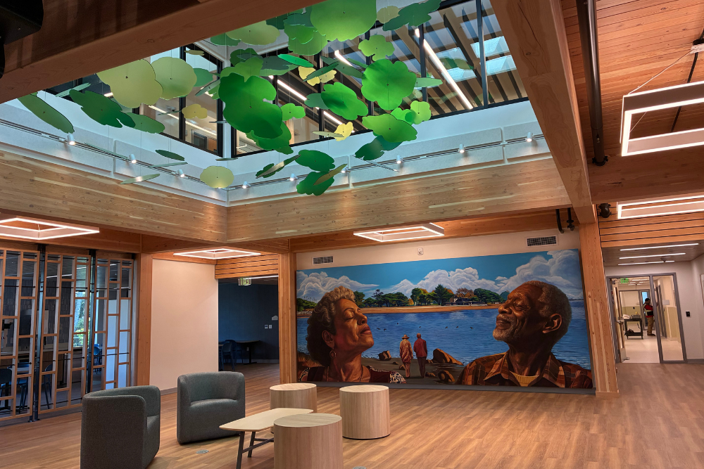 Bright indoor space with wood beams, hanging leaf art, and a large mural of two older adults by the water on the wall.