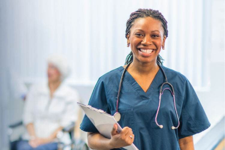 Healthcare worker in scrubs holding a clipboard and smiling, with a patient blurred in the background.