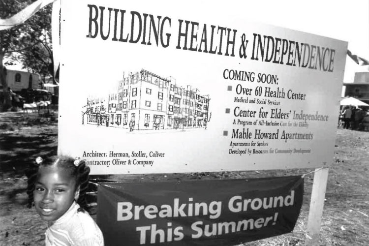Historic groundbreaking sign reading “Building Health & Independence” with development details and a child standing in the foreground.