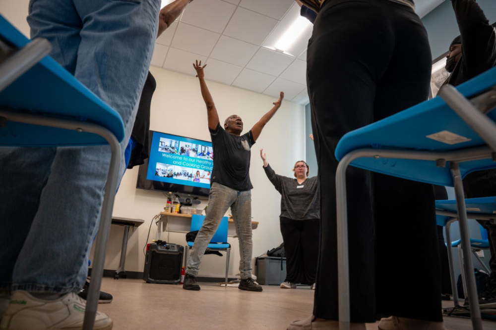 Participants take part in an interactive wellness or cooking group session, raising their hands during a guided activity.